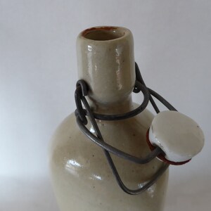 May include: A ceramic bottle with a metal wire closure and a white ceramic stopper. The bottle is a light beige color.
