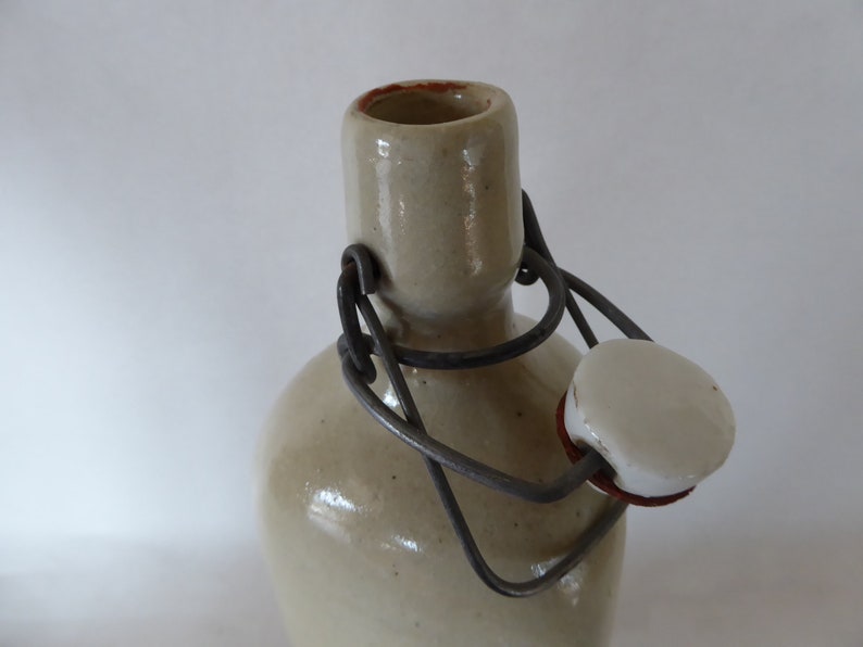 May include: A ceramic bottle with a metal wire closure and a white ceramic stopper. The bottle is a light beige color.
