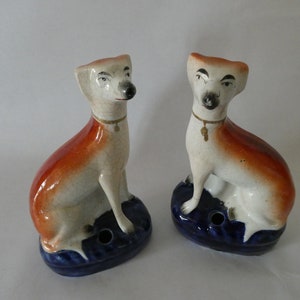 Pode incluir: Duas estatuetas de cães de cerâmica, cada uma com um design pintado em marrom e branco, sentadas em uma base azul. Os cães têm olhos pretos e uma coleira dourada com um pingente.