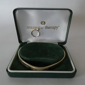 Op de afbeelding: Een groene fluwelen juwelendoos met een gouden armband en ring erin. Op de deksel van de doos staat "magnetic therapy" gedrukt.
