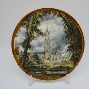 May include: Decorative plate with a gold rim featuring a detailed painting of a cathedral framed by trees. The artwork depicts a tall spire and surrounding landscape, with a cloudy sky backdrop.