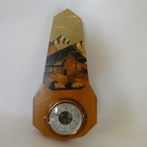 May include: A wooden barometer with a painted scene of a house in the mountains. The barometer has a round face with a needle and numbers indicating pressure.