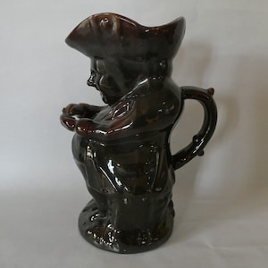 May include: A brown ceramic pitcher in the shape of a man wearing a hat. The pitcher has a handle and is glazed with a glossy finish. The figure is depicted with a jovial expression, holding his hands together.