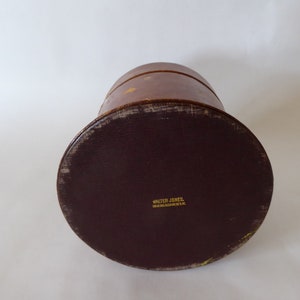 May include: A brown leather hat box with a dark brown leather bottom. The bottom has the text "Walter Jones,  Hat Manufacturer, U.S.A." printed in gold.