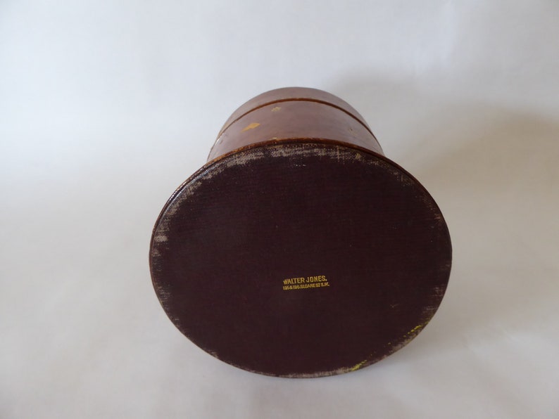 May include: A brown leather hat box with a dark brown leather bottom. The bottom has the text "Walter Jones,  Hat Manufacturer, U.S.A." printed in gold.