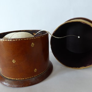 May include: A brown leather thread spool holder with gold accents and a white thread spool inside. The holder is open and the thread is partially pulled out.