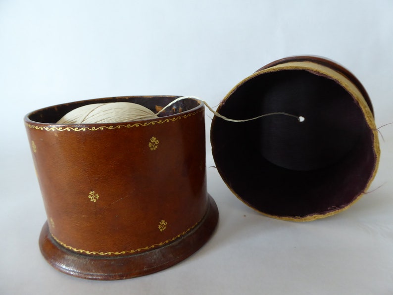May include: A brown leather thread spool holder with gold accents and a white thread spool inside. The holder is open and the thread is partially pulled out.