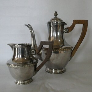 Pode incluir: Um conjunto de chá de prata com uma pega de madeira na chaleira e um jarro de creme de prata. A chaleira tem um design decorativo em torno da parte superior.