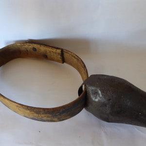 May include: A brown leather strap with a metal buckle attached to a large, dark gray metal cowbell.