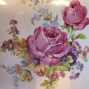 May include: A vintage floral design featuring pink and purple roses, yellow and white flowers, and green leaves on a white background.