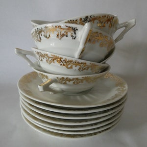 Pode incluir: Uma pilha de chávenas e pires de porcelana branca. As chávenas e os pires são decorados com padrões dourados. As chávenas têm pegas e estão empilhadas sobre os pires. Os pires estão empilhados.