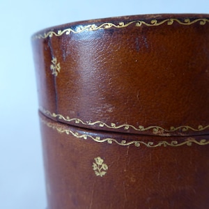 May include: A brown leather cylindrical box with gold trim and a floral design. The box has a lid and is made of high-quality leather.