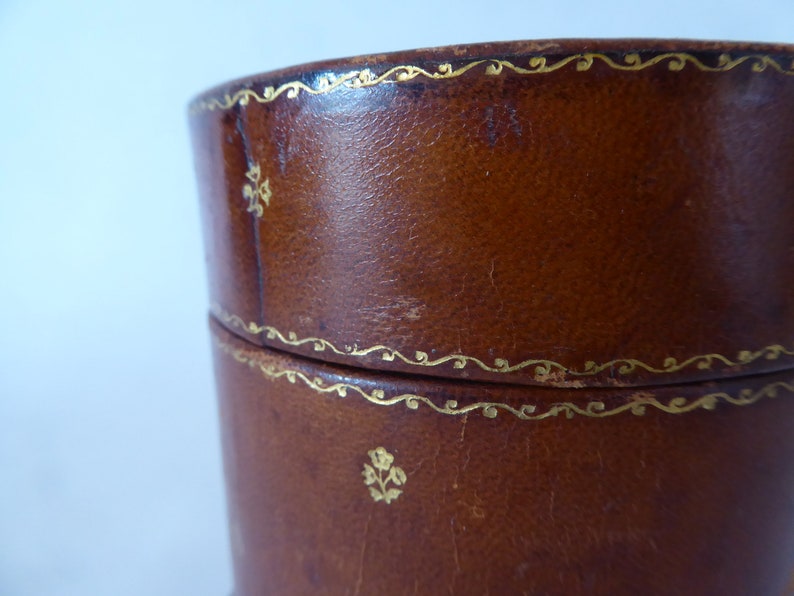 May include: A brown leather cylindrical box with gold trim and a floral design. The box has a lid and is made of high-quality leather.