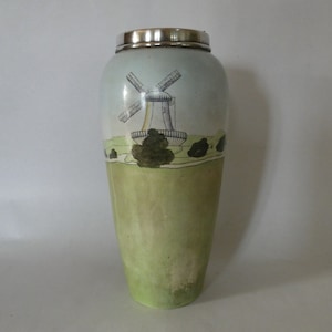 May include: A tall, green and white ceramic vase with a silver top. The vase features a hand-painted design of a windmill in a landscape.