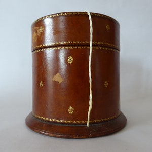 May include: A brown leather cylindrical box with gold accents and a white string attached to the lid. The box has a wooden base.