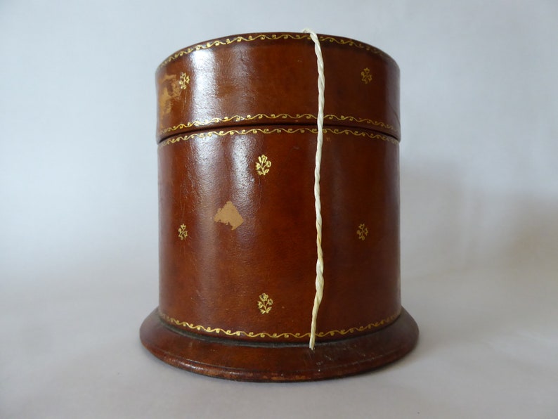 May include: A brown leather cylindrical box with gold accents and a white string attached to the lid. The box has a wooden base.