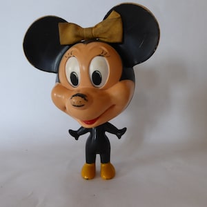 May include: Vintage Minnie Mouse figurine. The figure has a black body, large black ears, and a tan face. A yellow bow sits atop her head. Her shoes are yellow, and her mouth is red. The figurine is standing with arms outstretched.