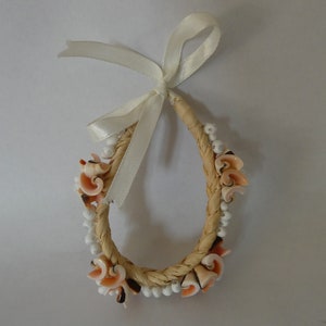 Può includere: Un braccialetto intrecciato di paglia con perline bianche e conchiglie rosa e marroni. Un nastro di raso bianco è legato attorno al braccialetto.