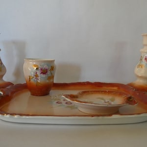 Pode incluir: Um conjunto de bandeja vintage de porcelana com dois castiçais, uma xícara pequena e um prato pequeno. O conjunto é decorado com desenhos florais em rosa, branco e dourado.