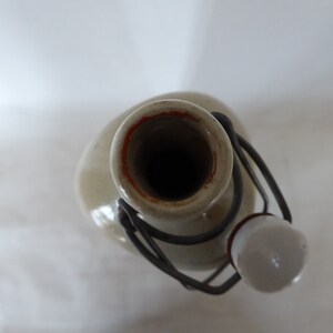 May include: A ceramic bottle with a metal lid and a white ceramic stopper. The bottle is a light gray color with a brown rim around the opening.