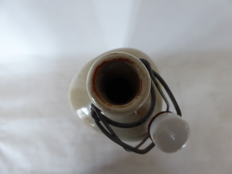 May include: A ceramic bottle with a metal lid and a white ceramic stopper. The bottle is a light gray color with a brown rim around the opening.