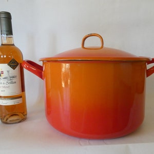Op de afbeelding: Een grote oranje en rode geëmailleerde pan met een deksel en twee handvatten. Naast de pan staat een fles wijn.