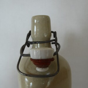 May include: A close-up of a ceramic bottle with a metal wire closure and a white ceramic stopper. The bottle is a light beige color and has a textured surface.