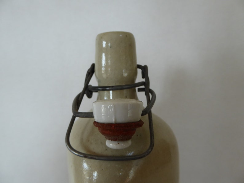 May include: A close-up of a ceramic bottle with a metal wire closure and a white ceramic stopper. The bottle is a light beige color and has a textured surface.