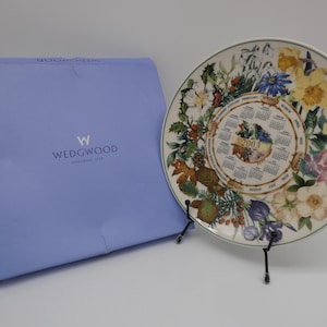 Może przedstawiać: Ozdobny talerz Wedgwood z kwiatowym wzorem i kalendarzem na rok 2003. Talerz jest prezentowany na czarnym stojaku, obok jasnoniebieskiego pudełka z logo Wedgwood.