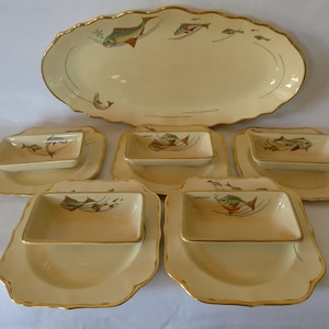 May include: A set of seven vintage ceramic serving dishes with a fish design. The dishes are cream colored with gold trim and feature a stylized fish design in shades of brown, green, and blue.