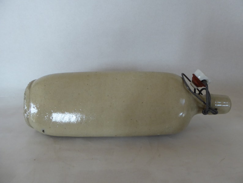 May include: A light beige ceramic bottle with a wire and ceramic stopper. The bottle has a rounded body and a narrow neck.