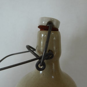 May include: A vintage stoneware bottle with a white ceramic lid and a metal wire handle. The bottle has a light brown glaze and a rustic appearance.