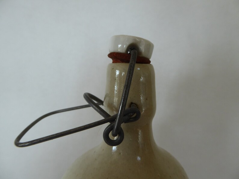 May include: A vintage stoneware bottle with a white ceramic lid and a metal wire handle. The bottle has a light brown glaze and a rustic appearance.