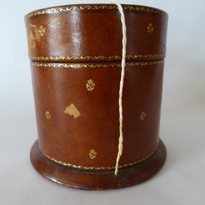 May include: A brown leather cylindrical box with gold trim and a white string hanging out of the top.