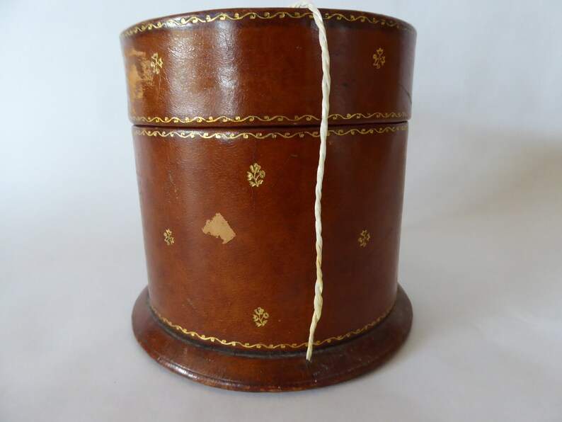 May include: A brown leather cylindrical box with gold trim and a white string hanging out of the top.