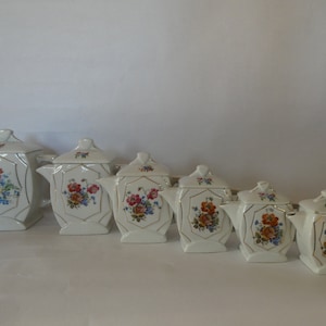 Könnte beinhalten: Satz von sechs weißen Porzellan-Teekannen mit Blumendesigns. Die Teekannen sind in einer Reihe vom größten zum kleinsten angeordnet. Jede Teekanne hat einen Deckel und einen Ausguss. Die Blumendesigns sind in den Farben Rosa, Rot, Gelb und Blau gehalten.