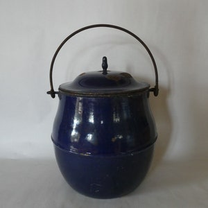 May include: A vintage blue enamel pot with a lid and a metal handle. The pot has a rounded shape and a dark blue color. The handle is arched and attached to the sides of the pot.
