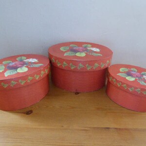 Puede incluir: Tres cajas ovaladas rojas con un diseño floral que presenta fresas y hojas. Las cajas están anidadas una dentro de la otra, con la caja más grande en la parte inferior y la caja más pequeña en la parte superior.