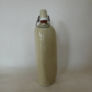 May include: A beige ceramic bottle with a metal swing top lid. The bottle has a textured surface and is slightly angled.