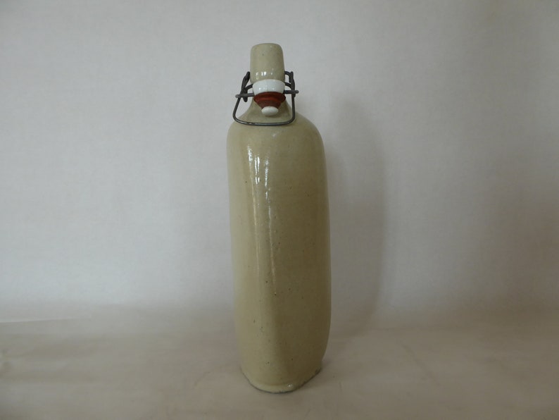 May include: A beige ceramic bottle with a metal swing top lid. The bottle has a textured surface and is slightly angled.
