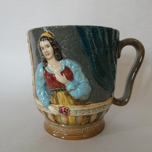 May include: A ceramic mug with a 3D design of a woman in a blue and red dress holding a rose. The mug has a handle and the words "Sweet Sorrow" on the bottom.