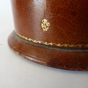 May include: Close-up of a brown leather box with a gold floral design and a gold trim around the top edge.