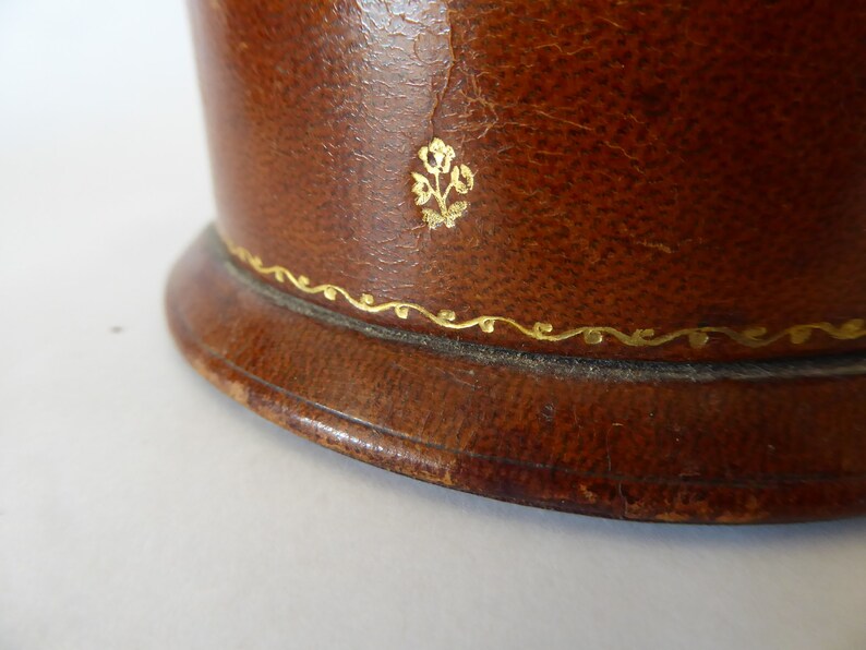 May include: Close-up of a brown leather box with a gold floral design and a gold trim around the top edge.