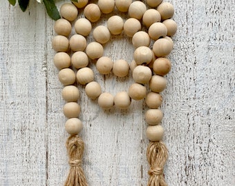 May include: A decorative wooden bead garland with two jute tassels. The natural wood beads are strung together in a looped pattern. The tassels are made of twisted jute fibers, adding a rustic touch. The garland is a neutral color.