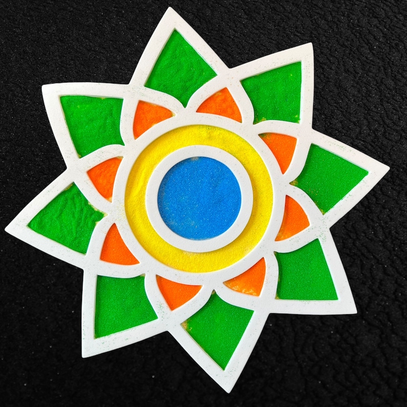 May include: A white star-shaped mandala with a blue, yellow, and orange center. The mandala is filled with green, orange, and yellow sand.