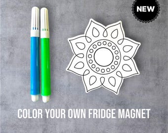 May include: A white, flower-shaped fridge magnet with a black outline, next to blue and green markers. The text "COLOR YOUR OWN FRIDGE MAGNET" is below the items. A black "NEW" sticker is in the upper right corner.
