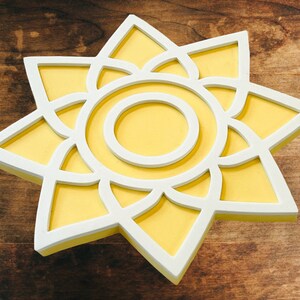 May include: A white and yellow sun-shaped wall decor with a geometric design. The sun has a white outline and a yellow center.