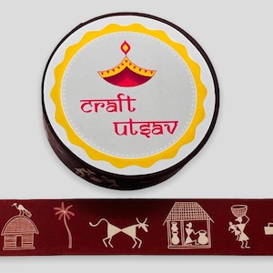 May include: A round, yellow and white sticker with a red and yellow diya design and the text "craft utsav". The sticker is on a brown background. Below the sticker is a red and white patterned ribbon with a design of a house, a tree, a cow, a woman carrying a basket, and a woman carrying a pot.