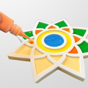 May include: A hand holds a bottle of orange glue and applies it to a white star-shaped craft. The star has a multi-colored design with green, orange, yellow, and blue sand.