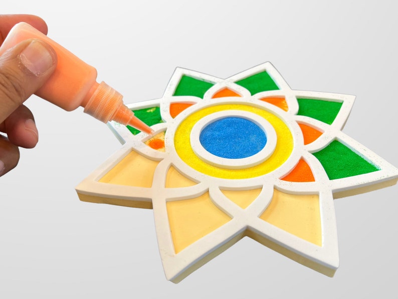 May include: A hand holds a bottle of orange glue and applies it to a white star-shaped craft. The star has a multi-colored design with green, orange, yellow, and blue sand.